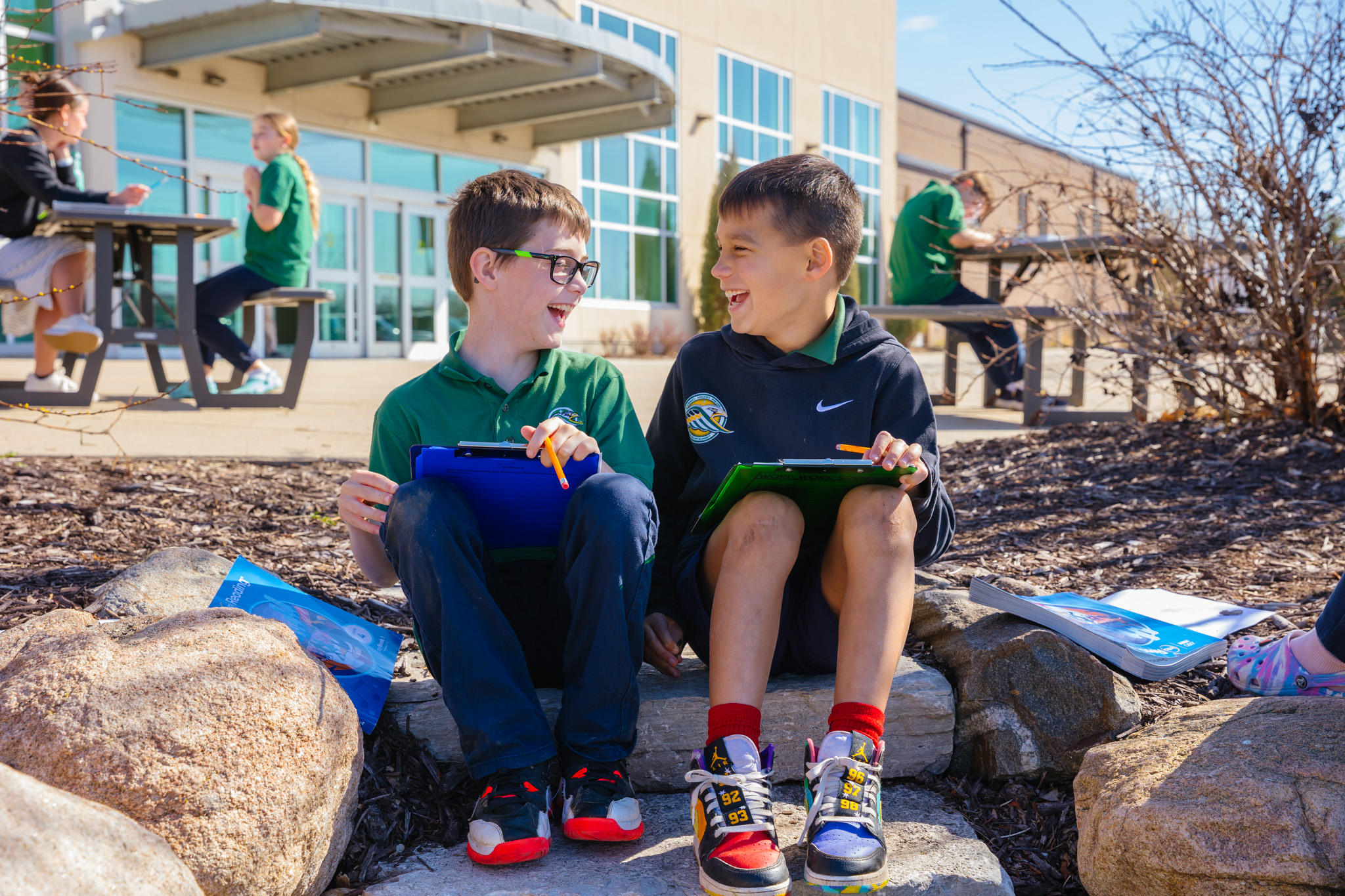 Two students sit on rock steps outside the front doors of the school working on their class assignment while giggling.
