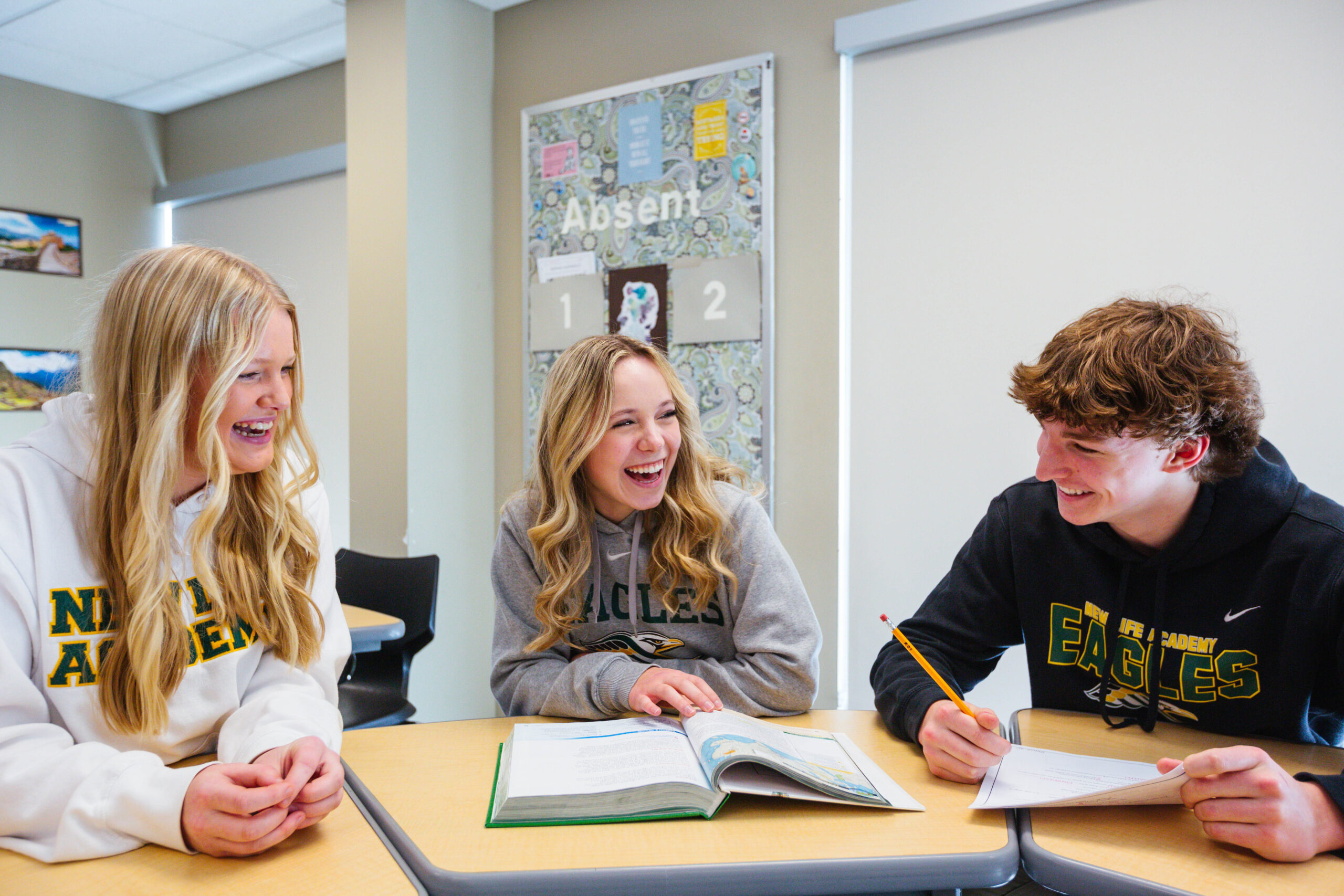 Three smiling students in New Life Academy sweatshirts are sitting at desks, engaged in a lively conversation during a classroom activity. They have open textbooks and papers in front of them.