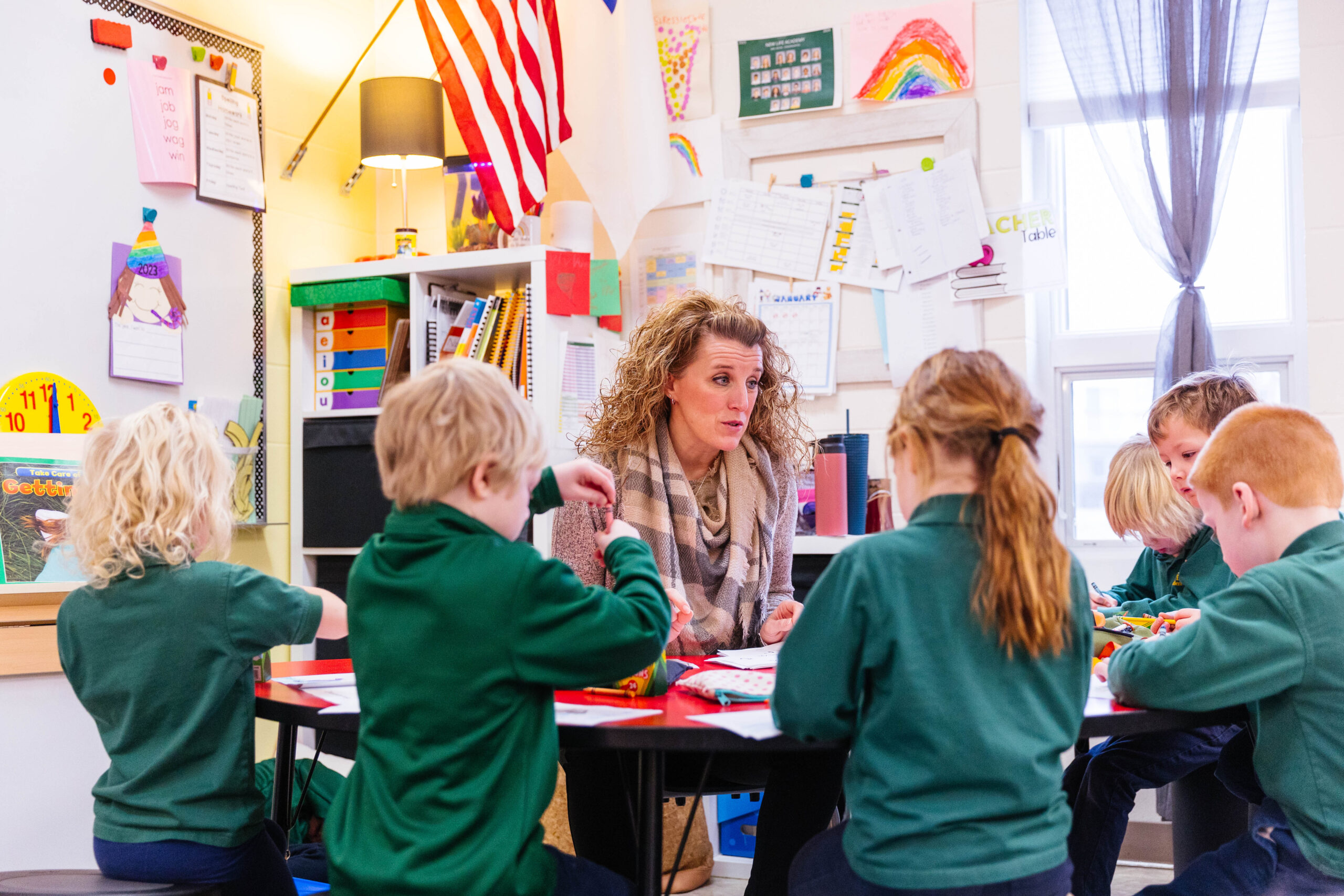 A kindergarten teacher at New Life Academy sits at a round table engaging with young students who are working on activities. The classroom is vibrant and colorful, with student artwork, educational materials, and an American flag displayed. The children are focused on their tasks, creating a lively and interactive learning environment.
