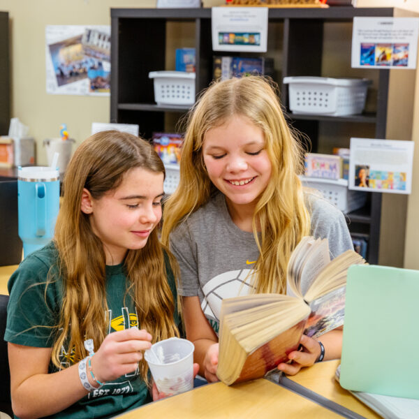 Two middle school girls on a swing outside hold their hands together to form a heart and smile at the camera.