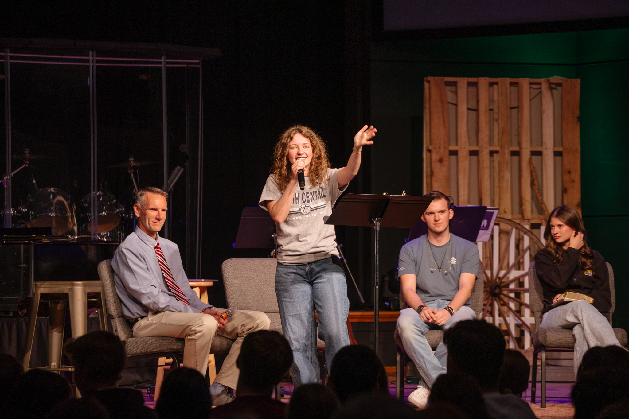 A high school student wearing a purple plaid shirt instructs students while standing in a classroom at New Life Academy. Other students are seated around tables, engaged in various activities such as cutting paper and studying, creating a lively and collaborative learning environment.