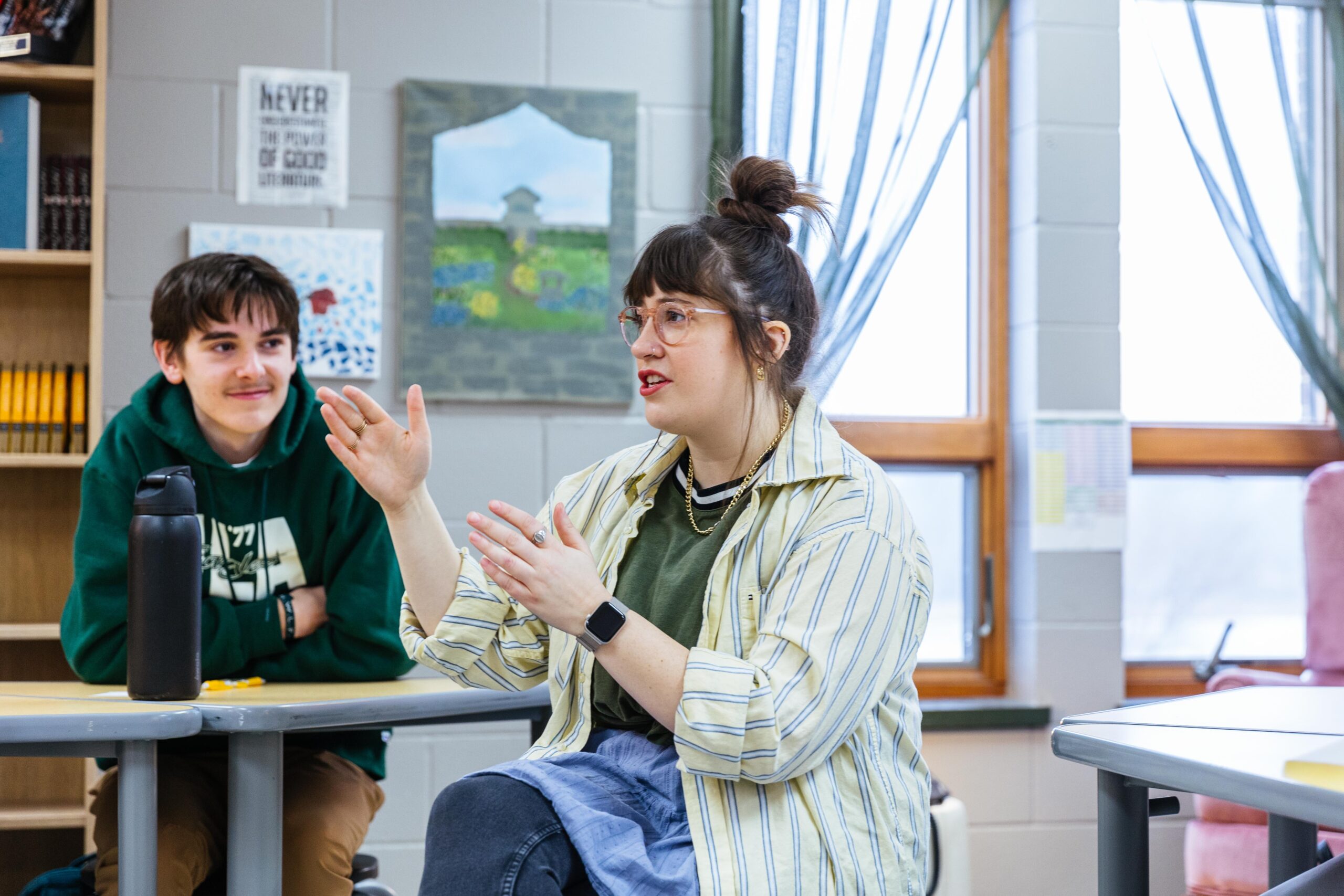 A high school student wearing a purple plaid shirt instructs students while standing in a classroom at New Life Academy. Other students are seated around tables, engaged in various activities such as cutting paper and studying, creating a lively and collaborative learning environment.