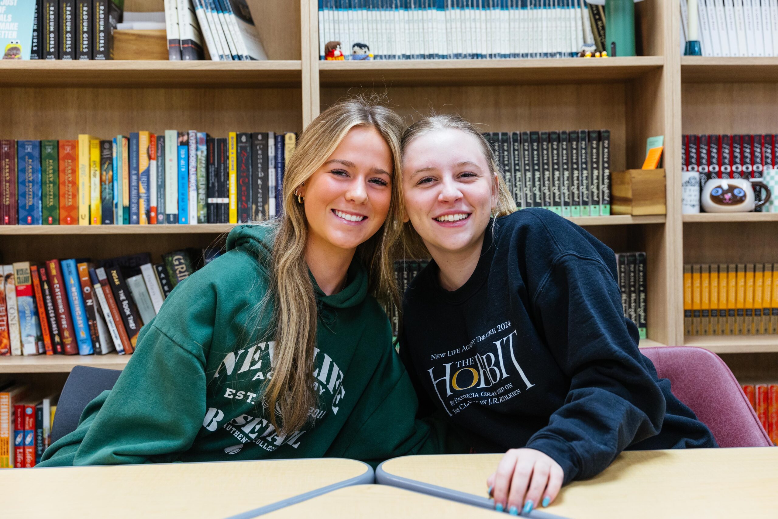A high school student wearing a purple plaid shirt instructs students while standing in a classroom at New Life Academy. Other students are seated around tables, engaged in various activities such as cutting paper and studying, creating a lively and collaborative learning environment.