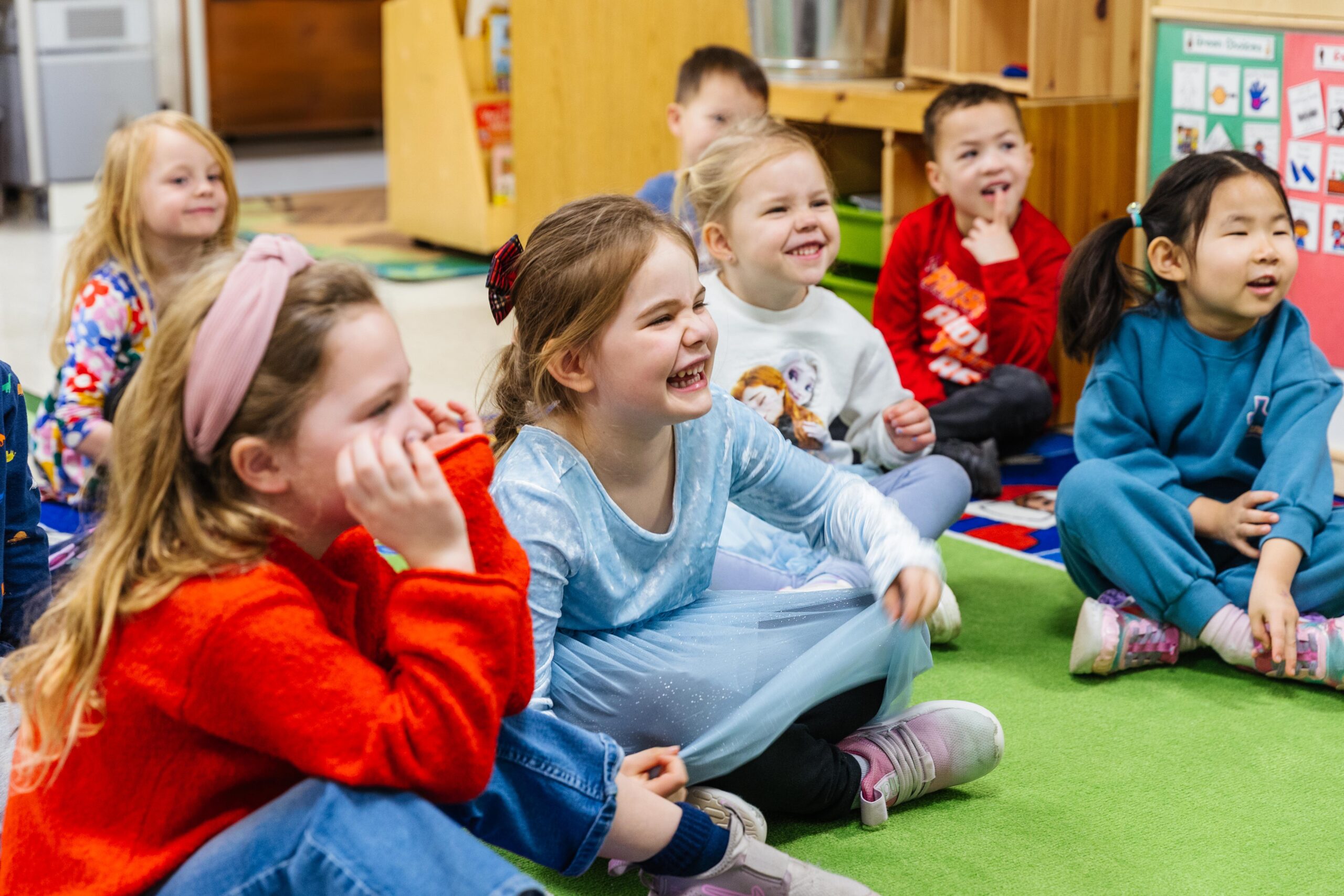 A teacher sits on a chair pointing to a colorful calendar on a classroom whiteboard while young children, sitting on a rainbow-colored carpet, raise their hands enthusiastically.