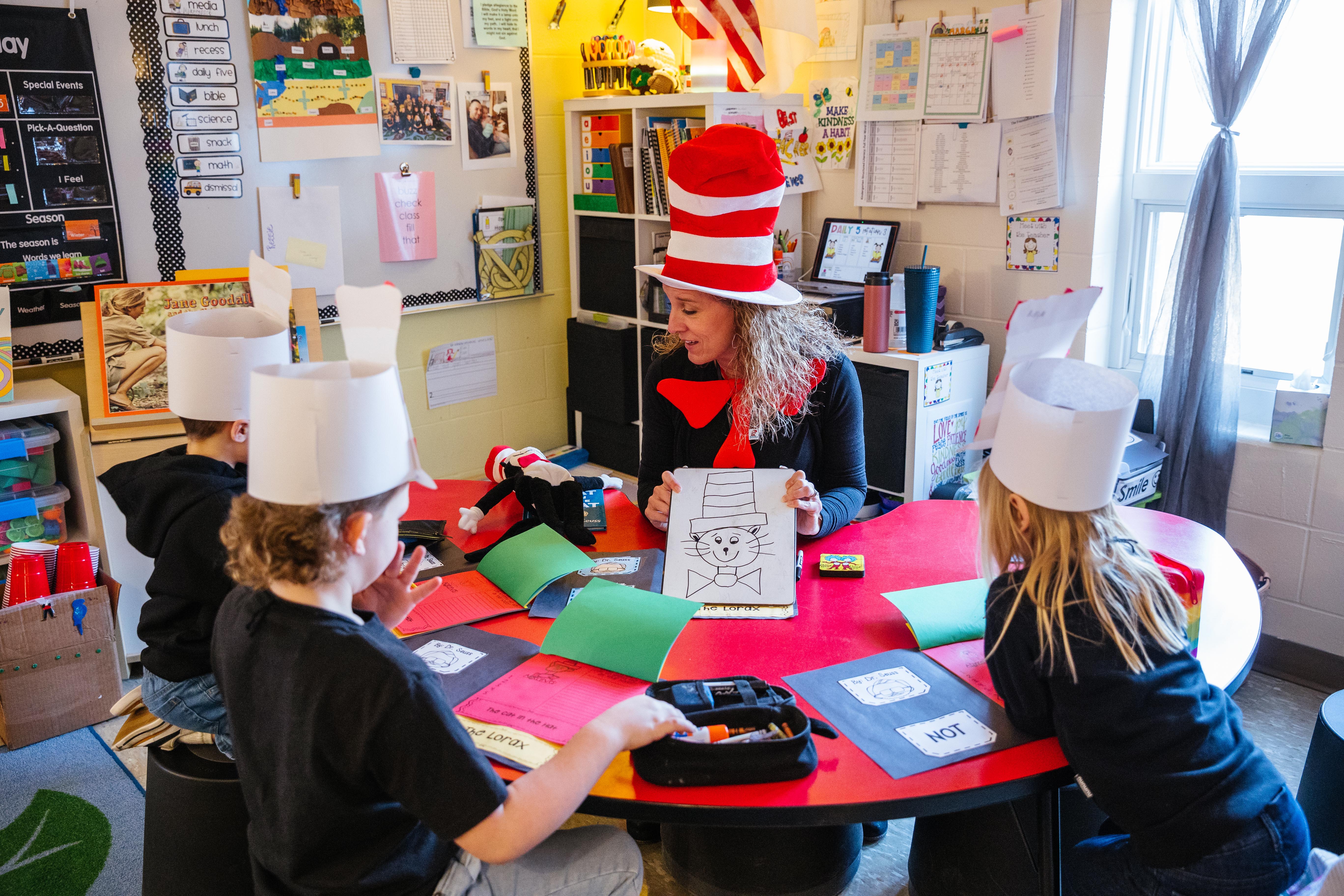 Kindergarten students sit around their classroom table learning from their teacher.