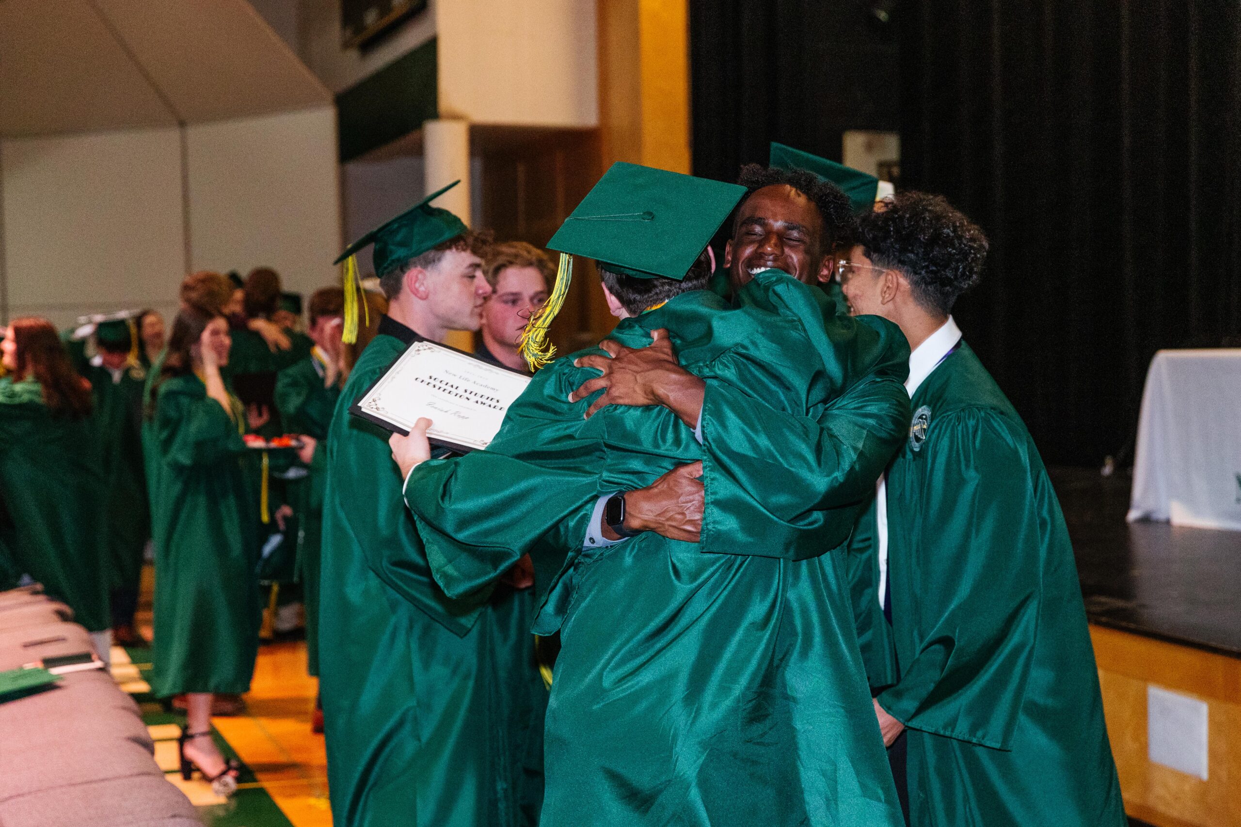 Three high school girls in New Life Academy volleyball jerseys stand outdoors, smiling and posing for a photo. They are wearing white jerseys with green and yellow accents, and the background features a sunny day with green trees and a lake.
