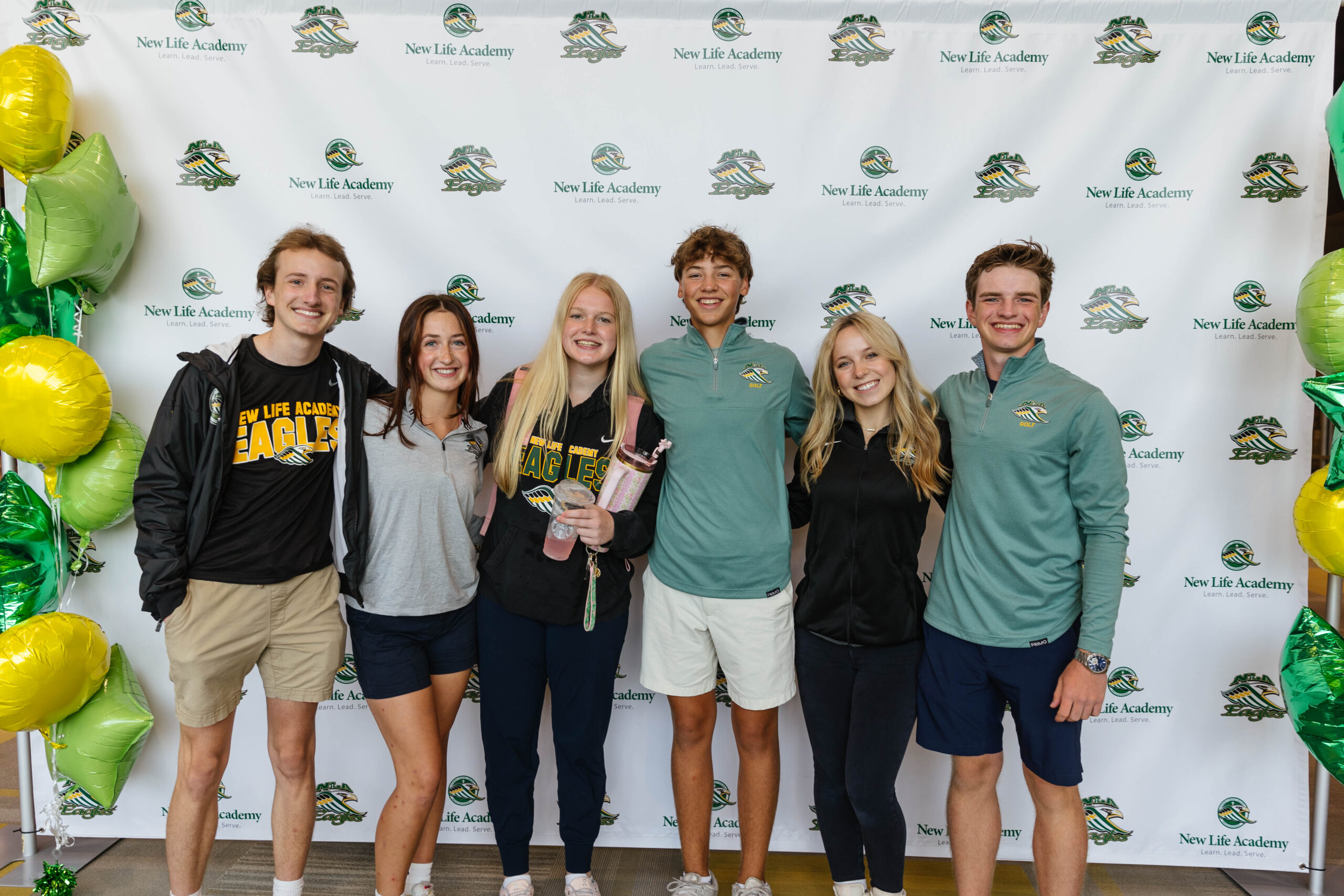 A group of high school students from New Life Academy's Student Council gather in a common area, wearing matching black hoodies that say "Student Council New Life Academy." They are smiling, chatting, and enjoying each other's company, with some seated on a couch and others standing around.