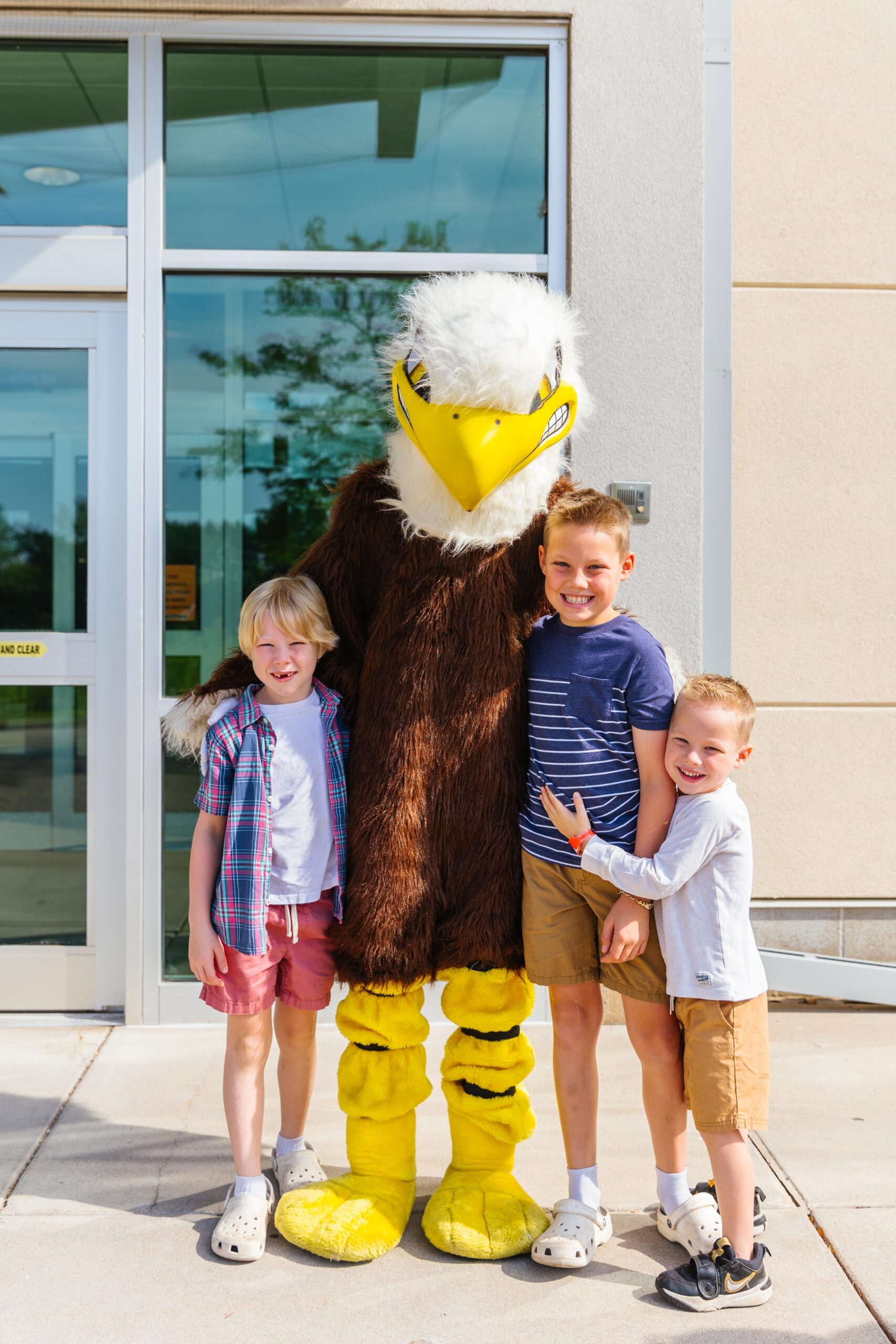 Soarin, New Life's Eagle Mascot, posing with two young boy students outside the main doors of NLA.
