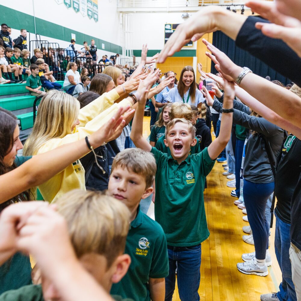Students at New Life Academy cheerfully walking through a tunnel of raised arms during a school spirit event in the gym.
