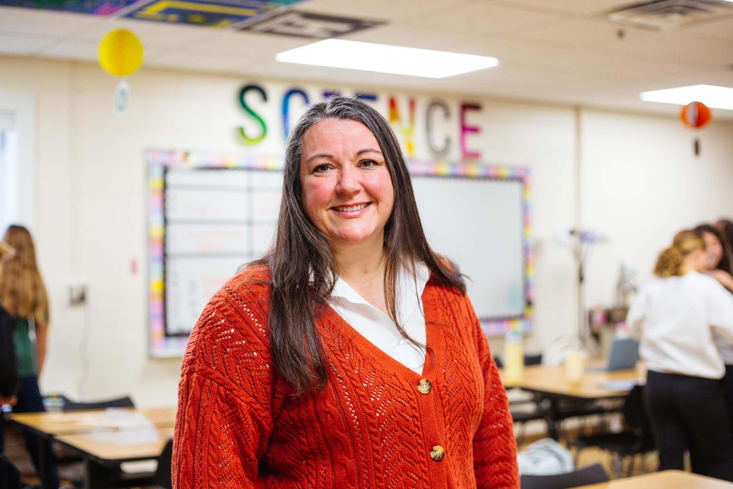 Smiling female science teacher wearing an orange sweater stands in a colorful classroom with students working in the background