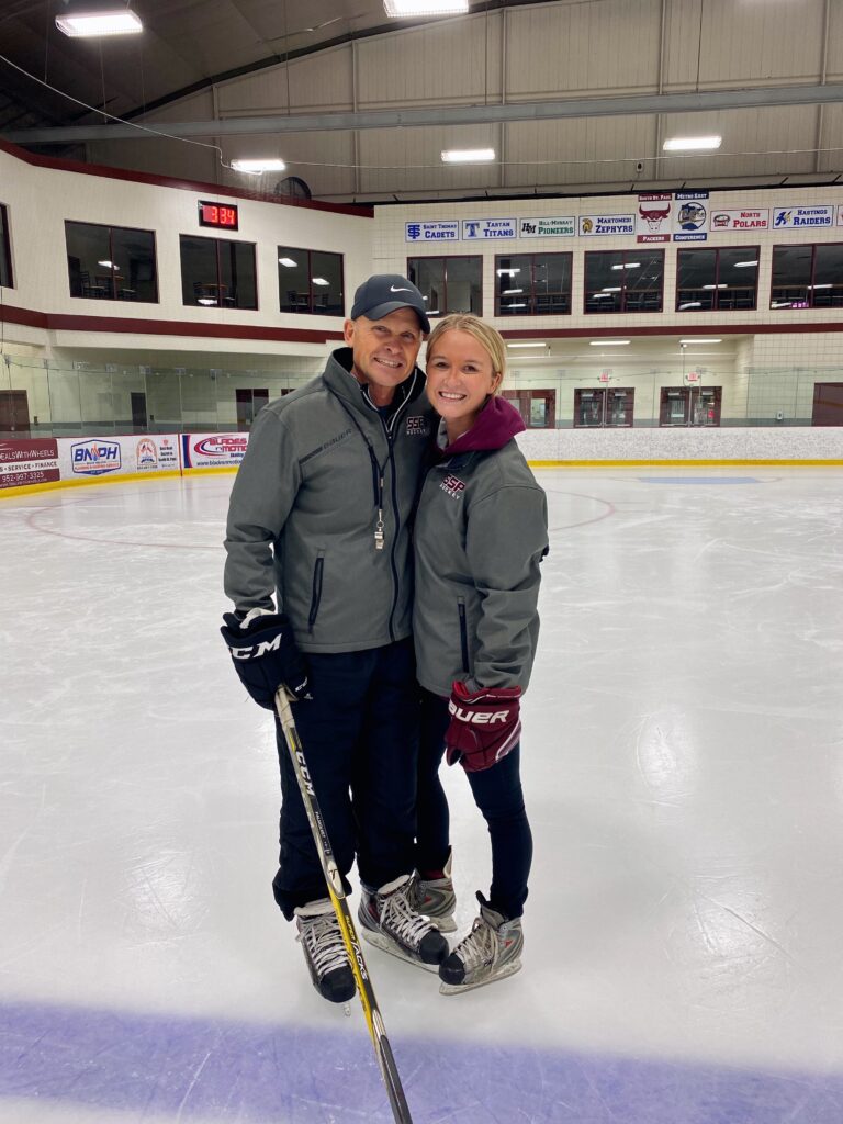 Father and daughter hockey coaches standing together on an ice rink, smiling and wearing skates and team jackets.