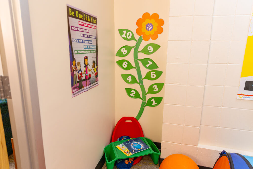 Corner of a sensory or reset room with a colorful flower number chart, motivational poster, small green table with books, and orange exercise ball.