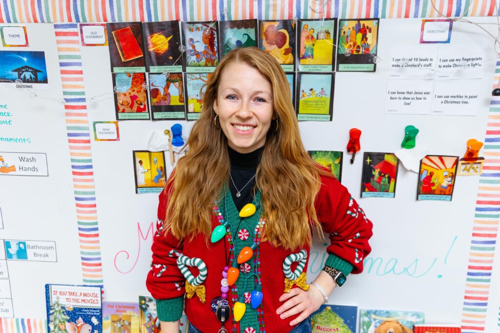 Teacher smiling in a classroom decorated with Bible story illustrations and Christmas decor.