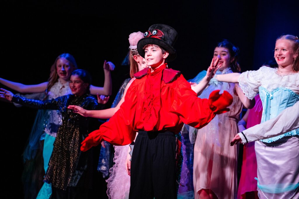 Student performers in a school production of The Little Mermaid Jr. with a cast member in a bright red costume leading the scene on stage.