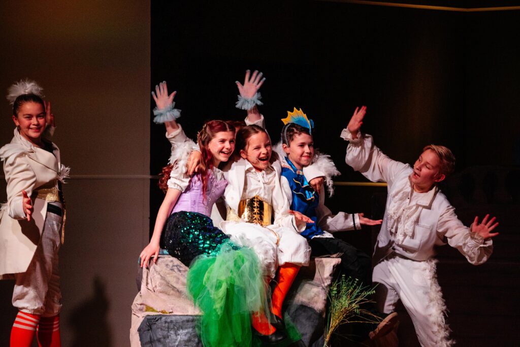 Children performing in a school production of The Little Mermaid Jr., smiling and posing together on stage while sitting on a rock set piece.