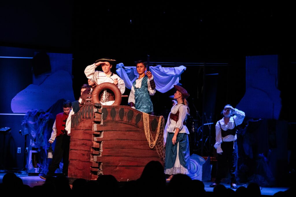 Middle school students performing a scene from The Little Mermaid Jr. on stage, featuring a ship set with nautical props and costumes.