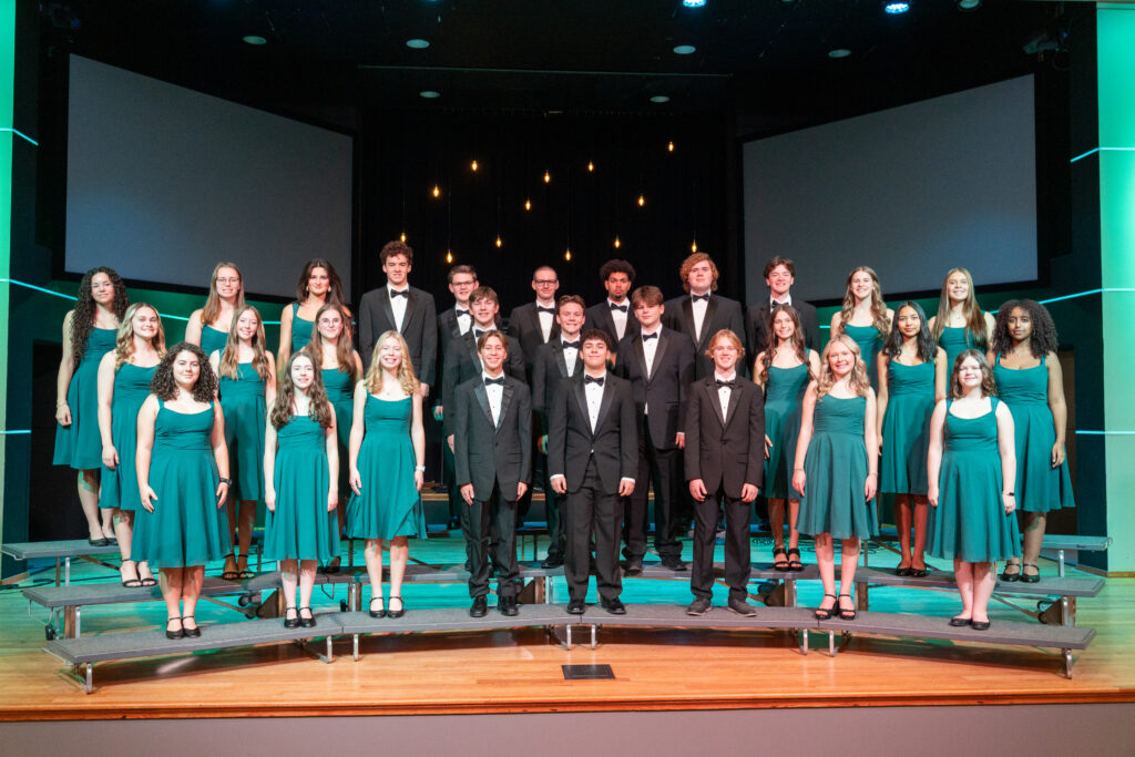 Morningstar Choir at New Life Academy posed on stage in formal green dresses and black tuxedos for a group photo.