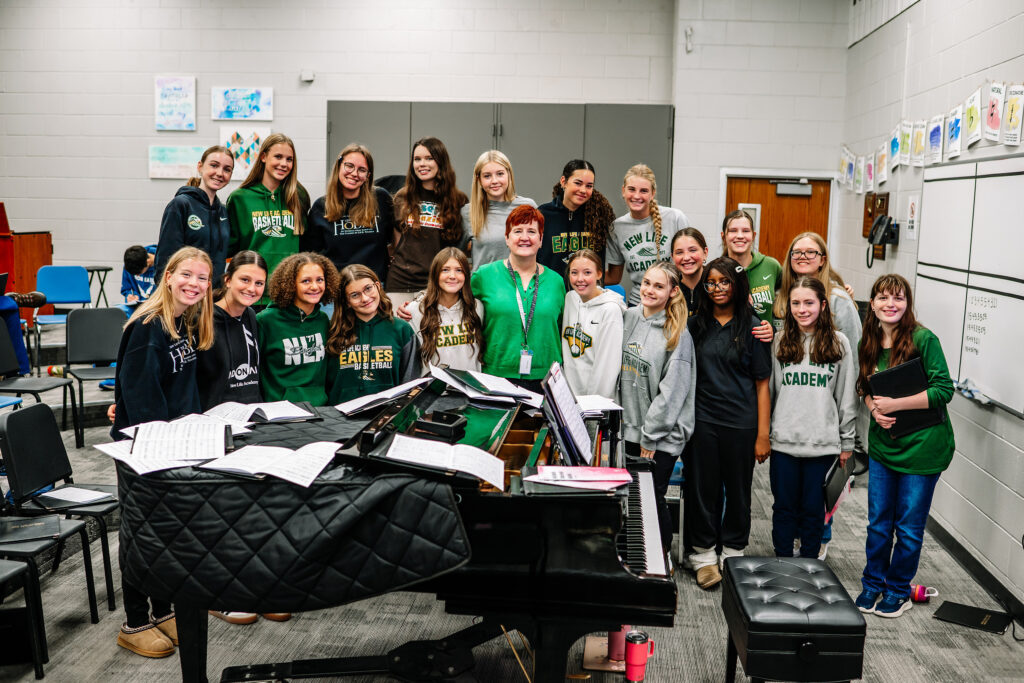 Group of choir students standing together in a music classroom at New Life Academy with a piano in the foreground.