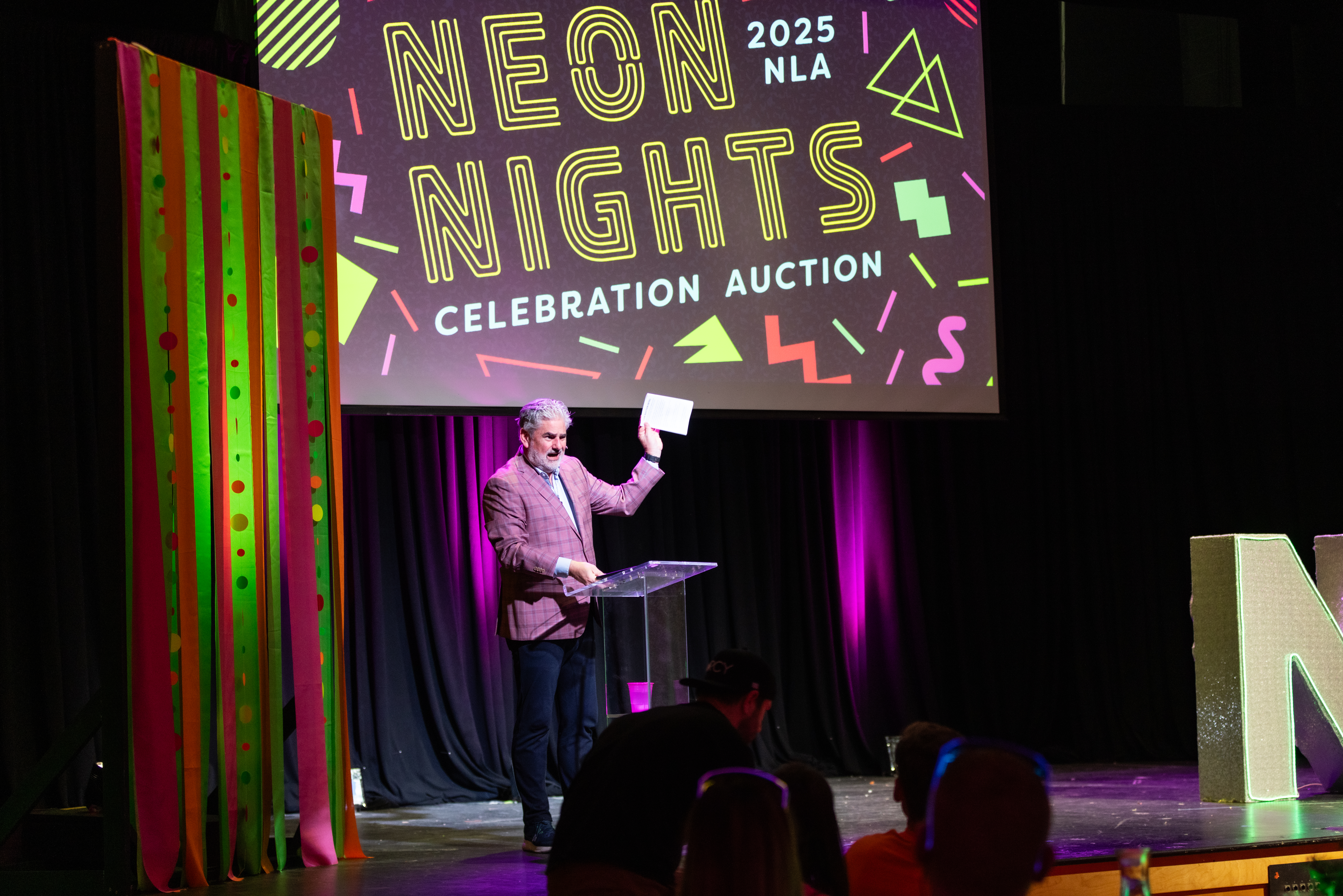 Stage at Neon Nights Celebration Auction with colorful neon-themed backdrop and large illuminated letters, part of New Life Academy’s annual fundraising gala.
