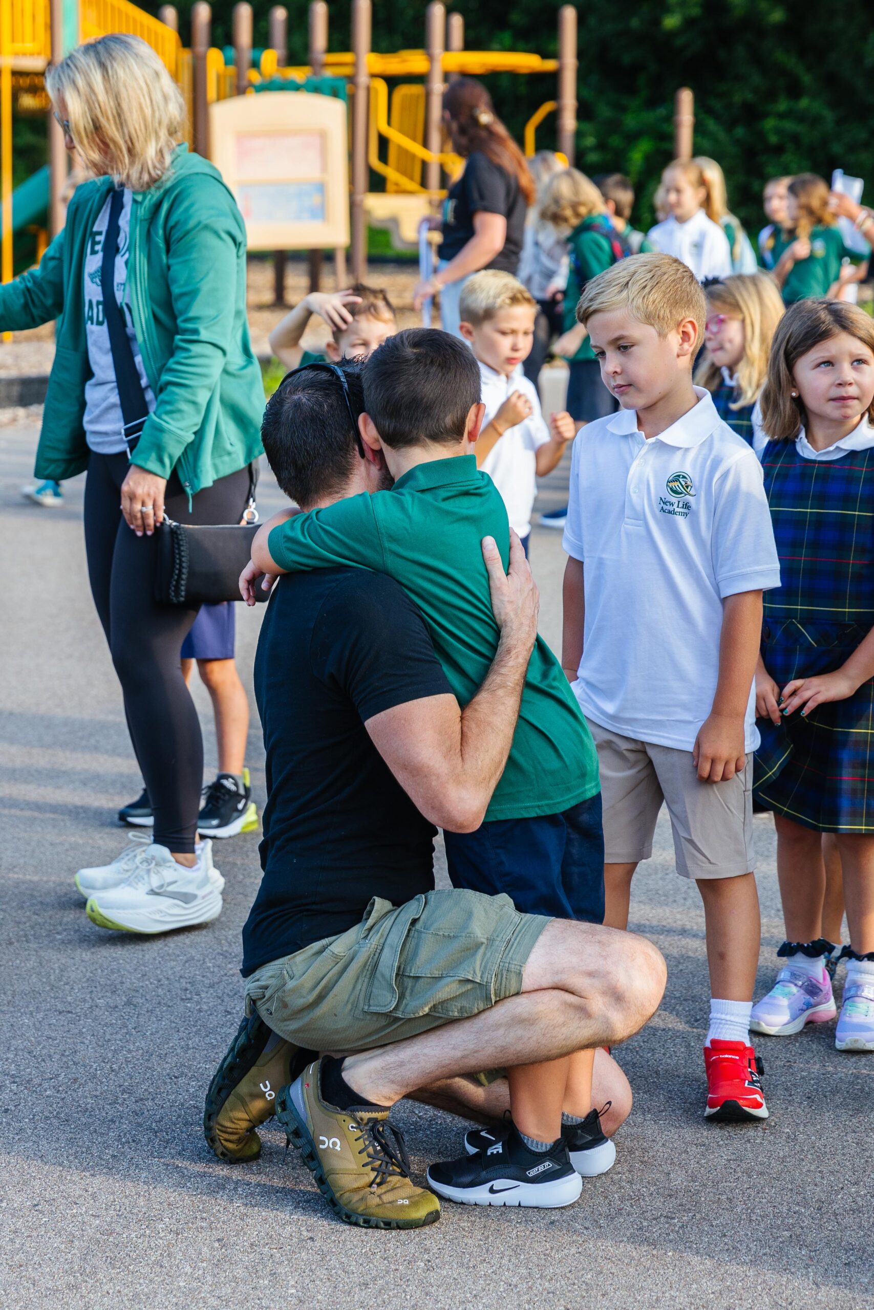 A high school student wearing a purple plaid shirt instructs students while standing in a classroom at New Life Academy. Other students are seated around tables, engaged in various activities such as cutting paper and studying, creating a lively and collaborative learning environment.
