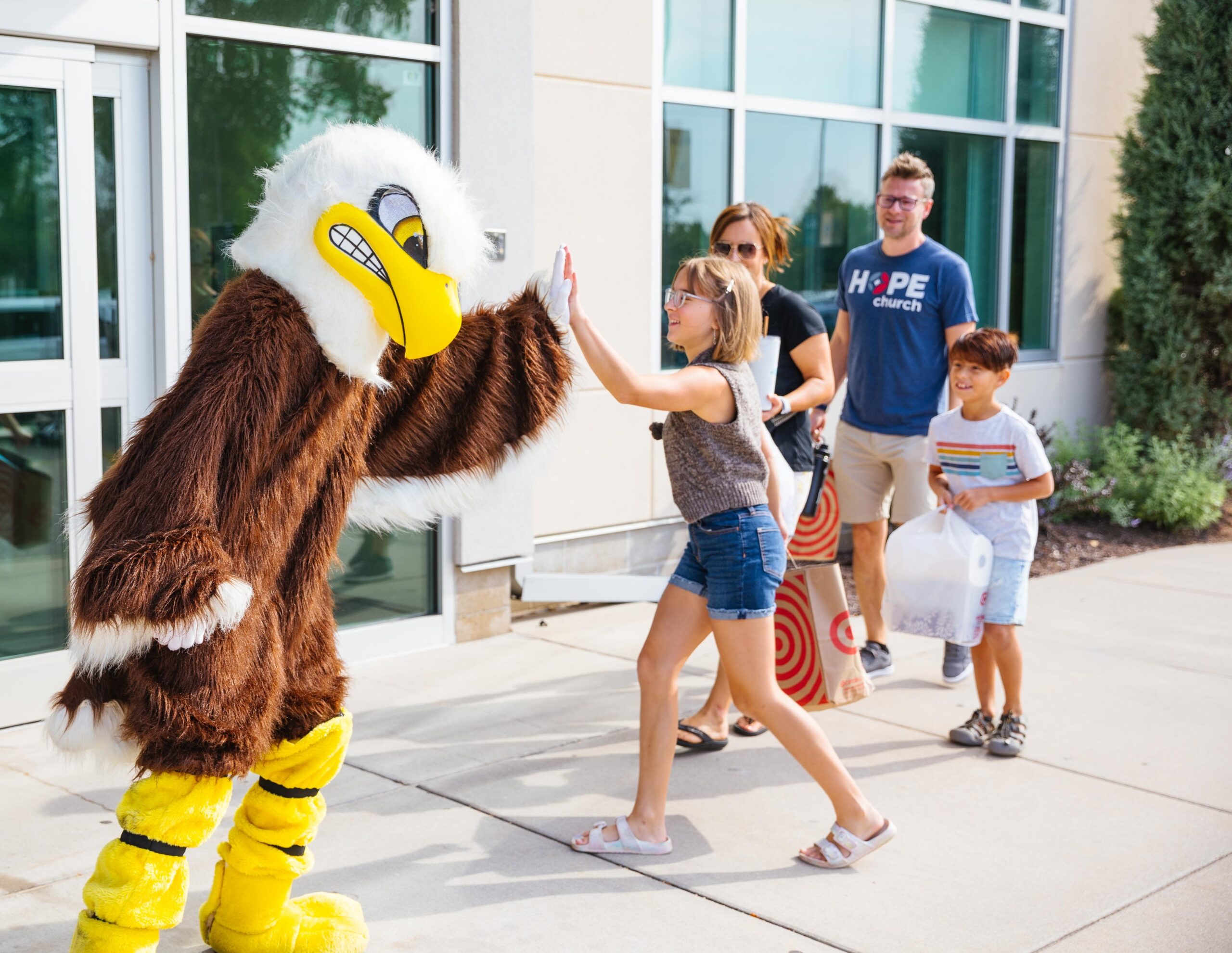 A high school student wearing a purple plaid shirt instructs students while standing in a classroom at New Life Academy. Other students are seated around tables, engaged in various activities such as cutting paper and studying, creating a lively and collaborative learning environment.