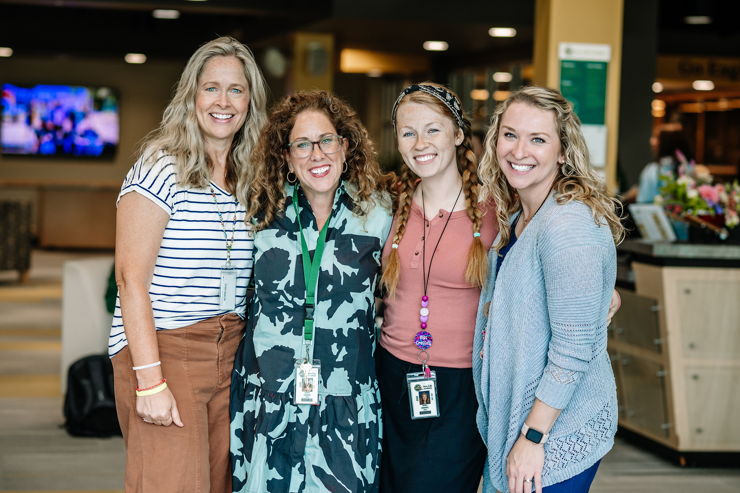 Three smiling students in New Life Academy sweatshirts are sitting at desks, engaged in a lively conversation during a classroom activity. They have open textbooks and papers in front of them.