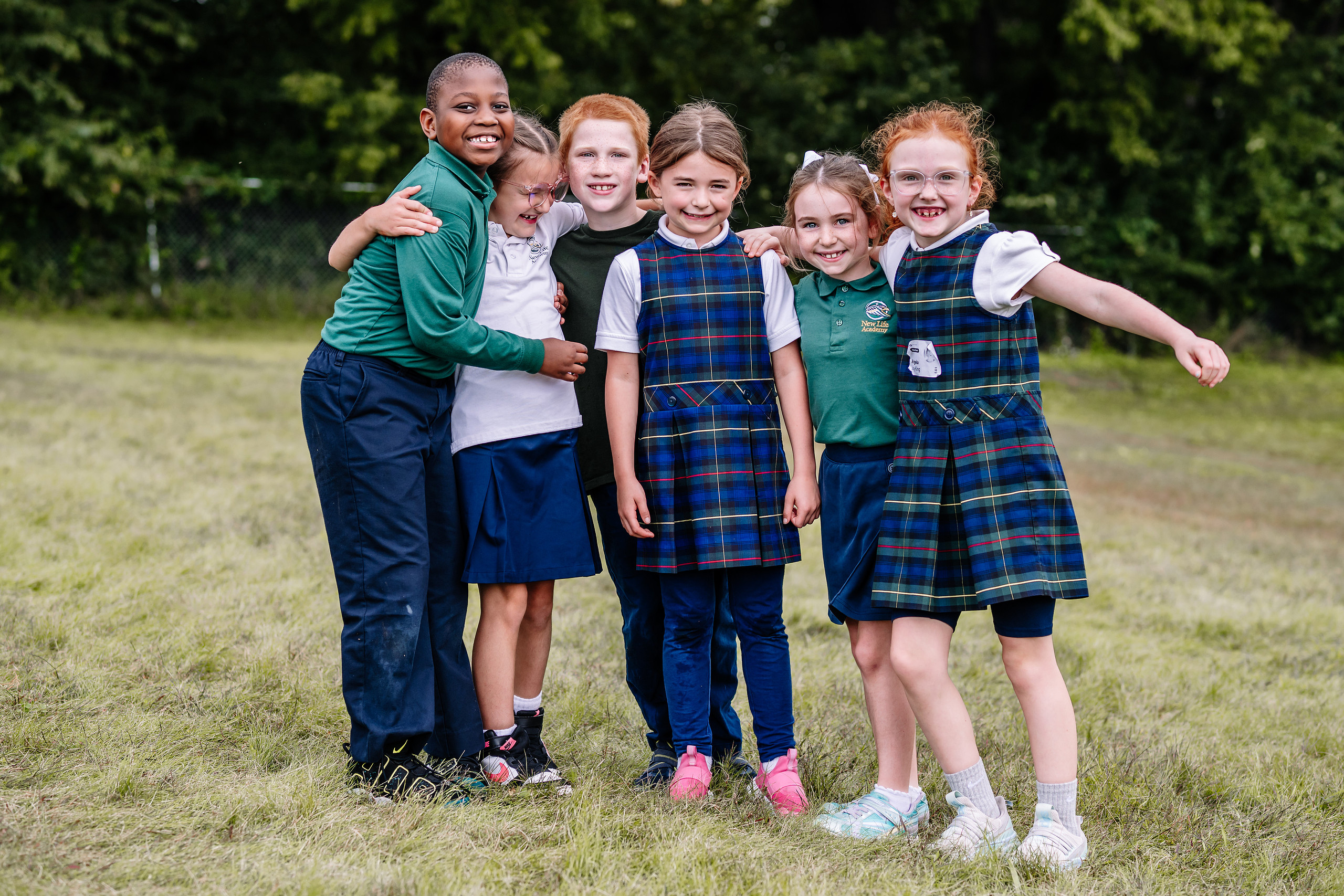 Three smiling students in New Life Academy sweatshirts are sitting at desks, engaged in a lively conversation during a classroom activity. They have open textbooks and papers in front of them.