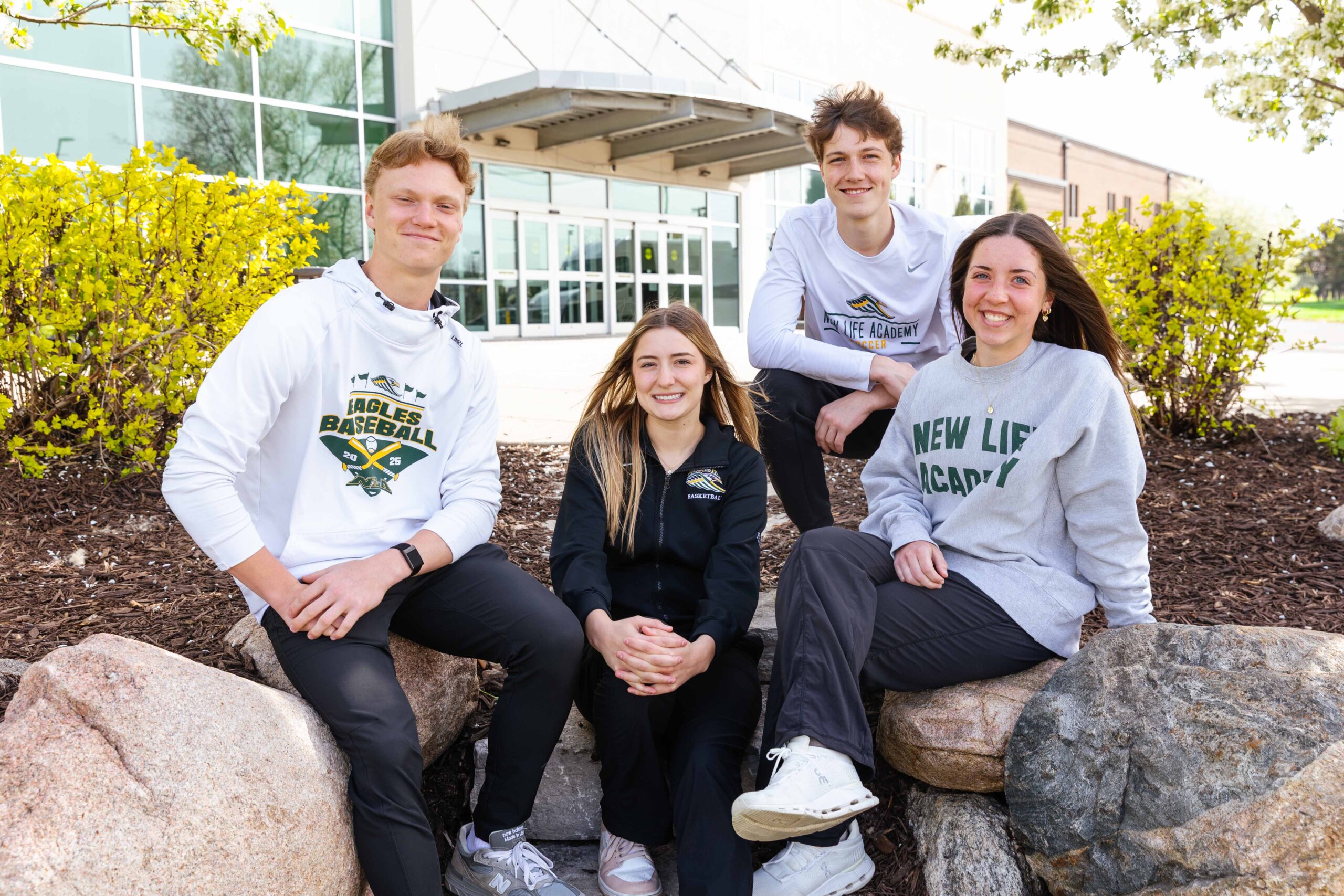 Three smiling students in New Life Academy sweatshirts are sitting at desks, engaged in a lively conversation during a classroom activity. They have open textbooks and papers in front of them.